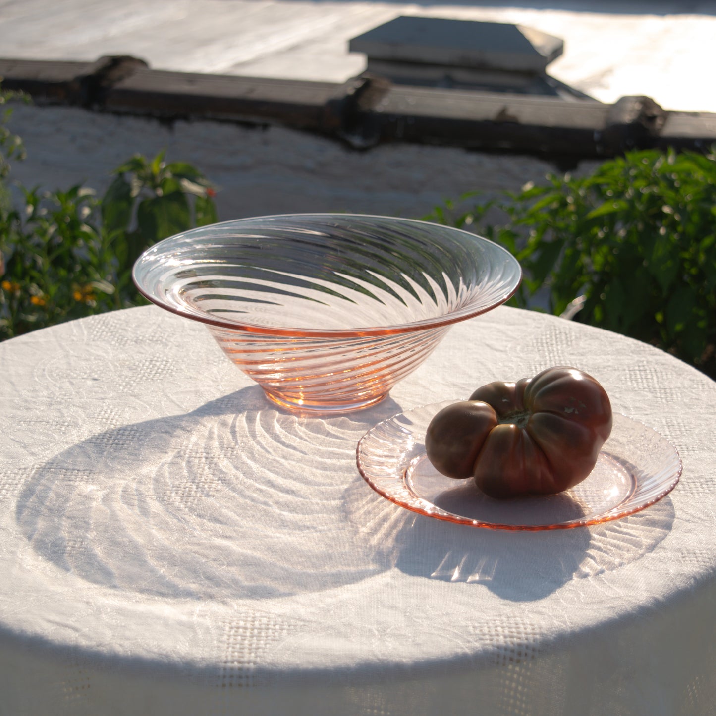 Pink glass bowl next to an heirloom tomato on a pink plate, all on a white lace tablecloth with a blurred garden background